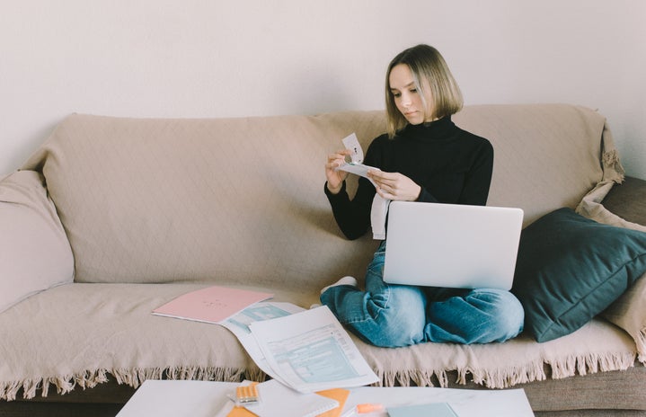 Woman with chin-length hair and a black turtleneck sits on a couch with her laptop and papers.