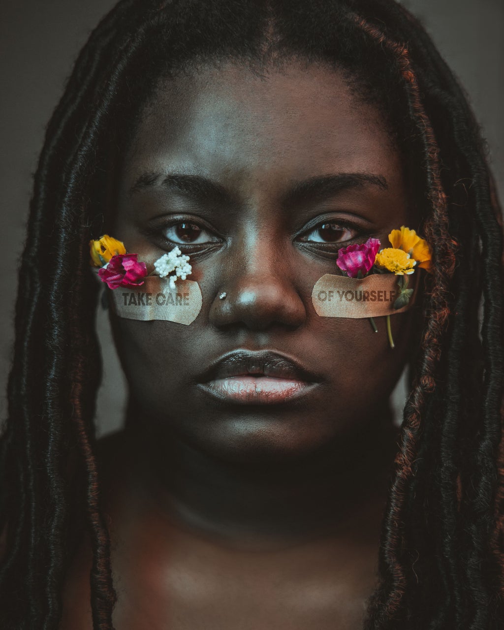Black woman with yellow and pink flowers taped to her face with  \'Take care of yourself\' written on the tape