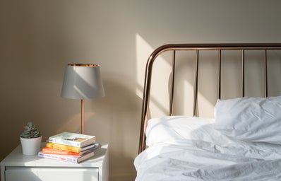 The image is focused on a desk. The desk has a lamp situated on it, with a stack of books and a fake plant as well. There is a partial view of a bed with white sheets.