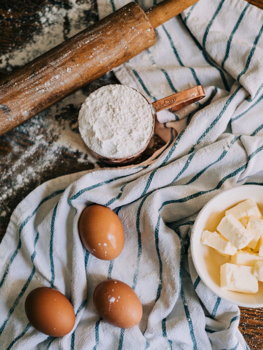 Brown eggs, butter in a bowl, and flour on a striped dish towel