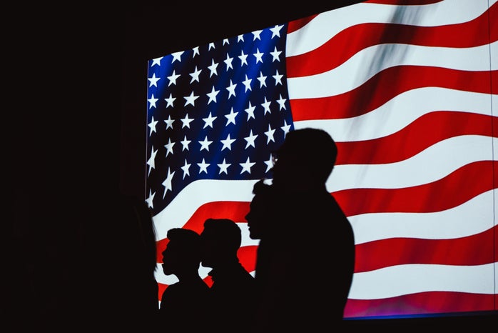 Four silhouettes stand in front of a digital American flag