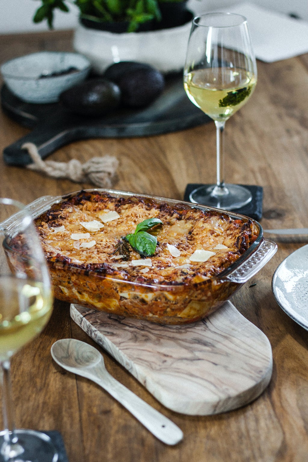 lasagna on a wooden table surrounded by wooden serving spoon and glasses of white wine