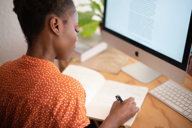 Black woman writing at computer desk writing in journal notebook write natural work corporate african