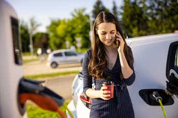 Young woman talking on mobile phone while waiting for electric car to charge