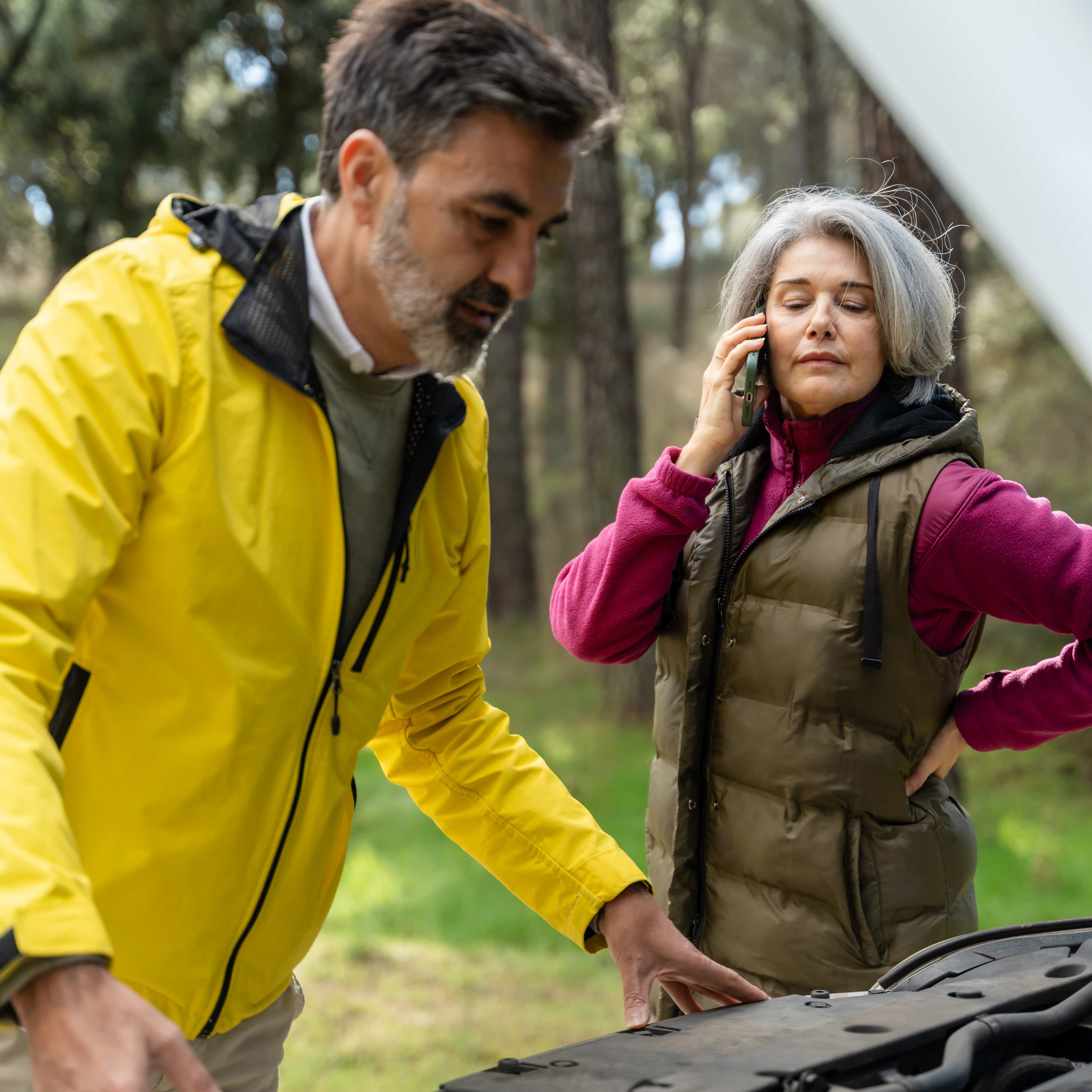 Mature couple is having car trouble in the forest, examining the engine and calling for assistance