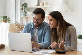 Happy young couple husband and wife using laptop computer looking at screen pay bills online in app calculate mortgage investment payment