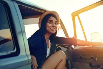Smiling young woman getting out of a car against a sunset