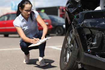 Woman inspector writing in documents on clipboard near broken car.