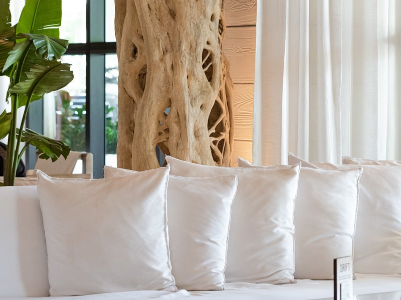 Five white pillows line an equally white sofa.  A driftwood style art installation compliments the couch as a backdrop piece.