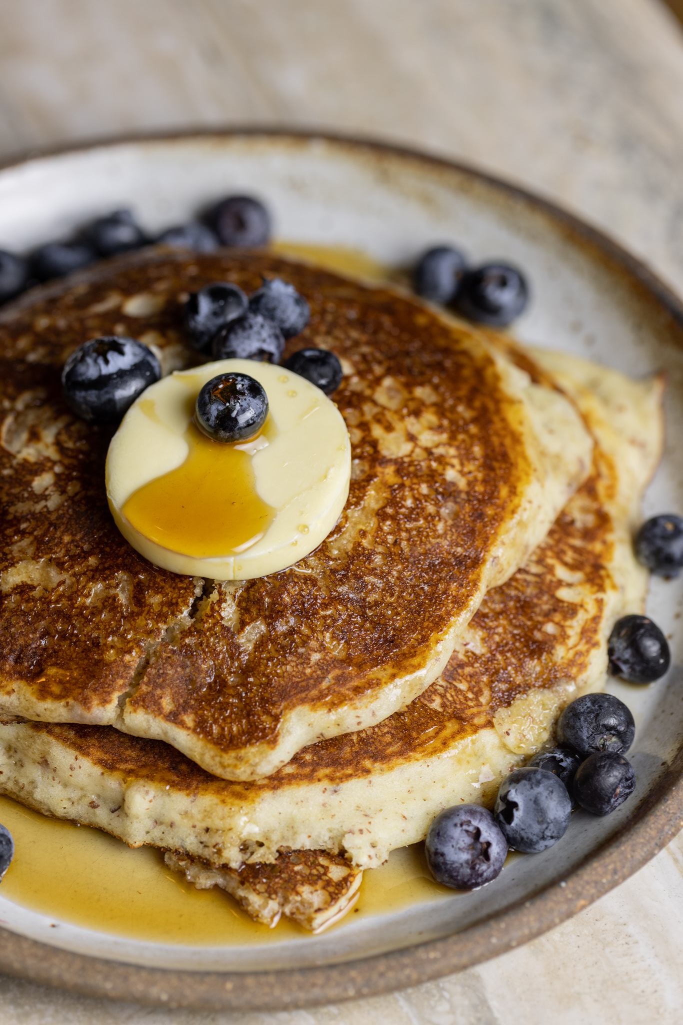 A stack of blueberry pancakes with butter and syrup poured ontop.  Blueberries cover the pancakes and plate.