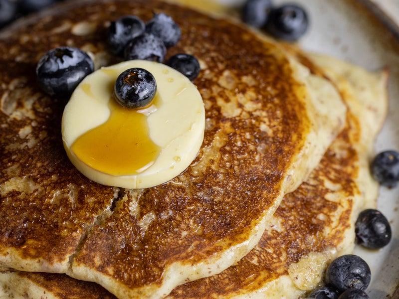 A stack of blueberry pancakes with butter and syrup poured ontop.  Blueberries cover the pancakes and plate.