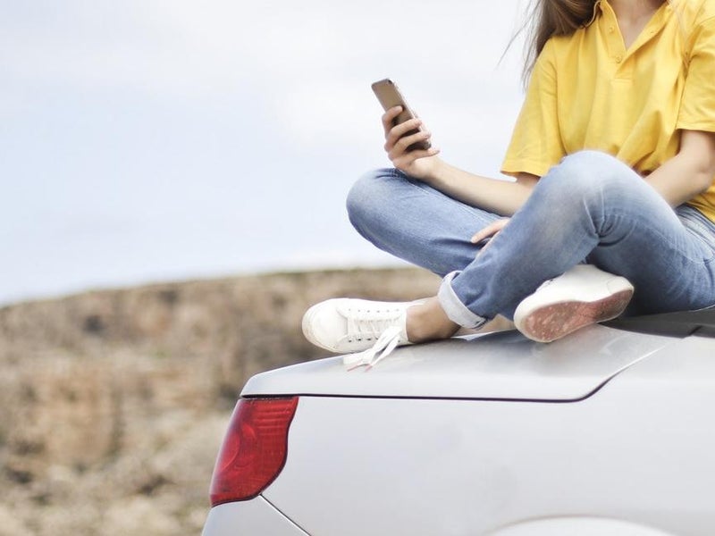 A woman wearing a yellow polo and faded blue jeans sits cross legged atop a white car with its top dropped back