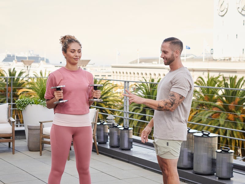 A male private trainer instructs a female client holding free weights, on the rooftop of 1 Hotels SFO