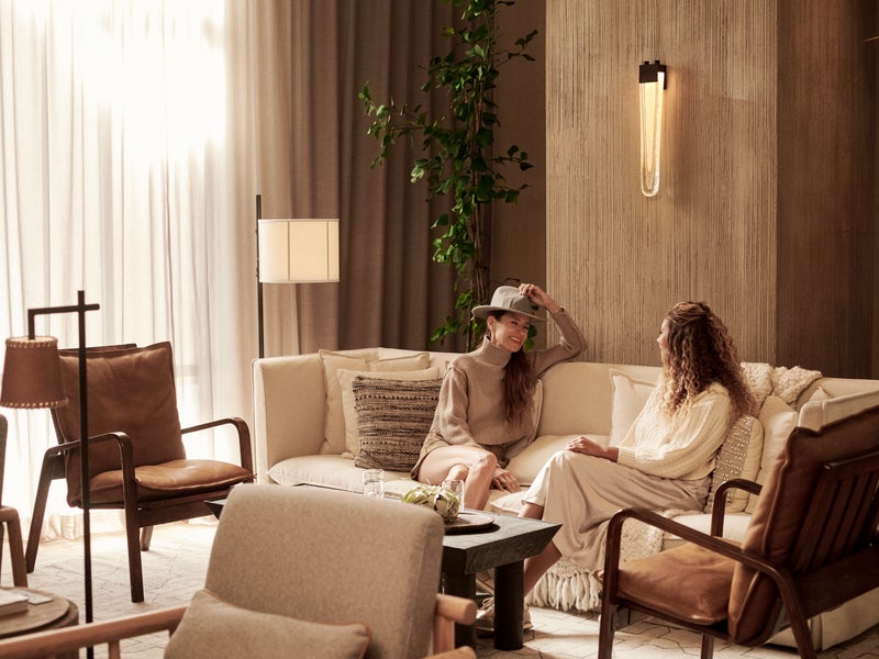 Two females dressed in white and beige sit on a white couch in the 1 Hotels SFO lobby area