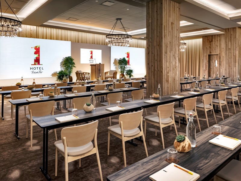 Black wooden tables stretch lengthwise across a 1 Hotels meeting room. Three projector screens line the wall in which all seats are facing.