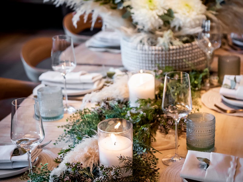 A wooden style banquet table decorated with white and green flowers, lights, and an elegant table spread.