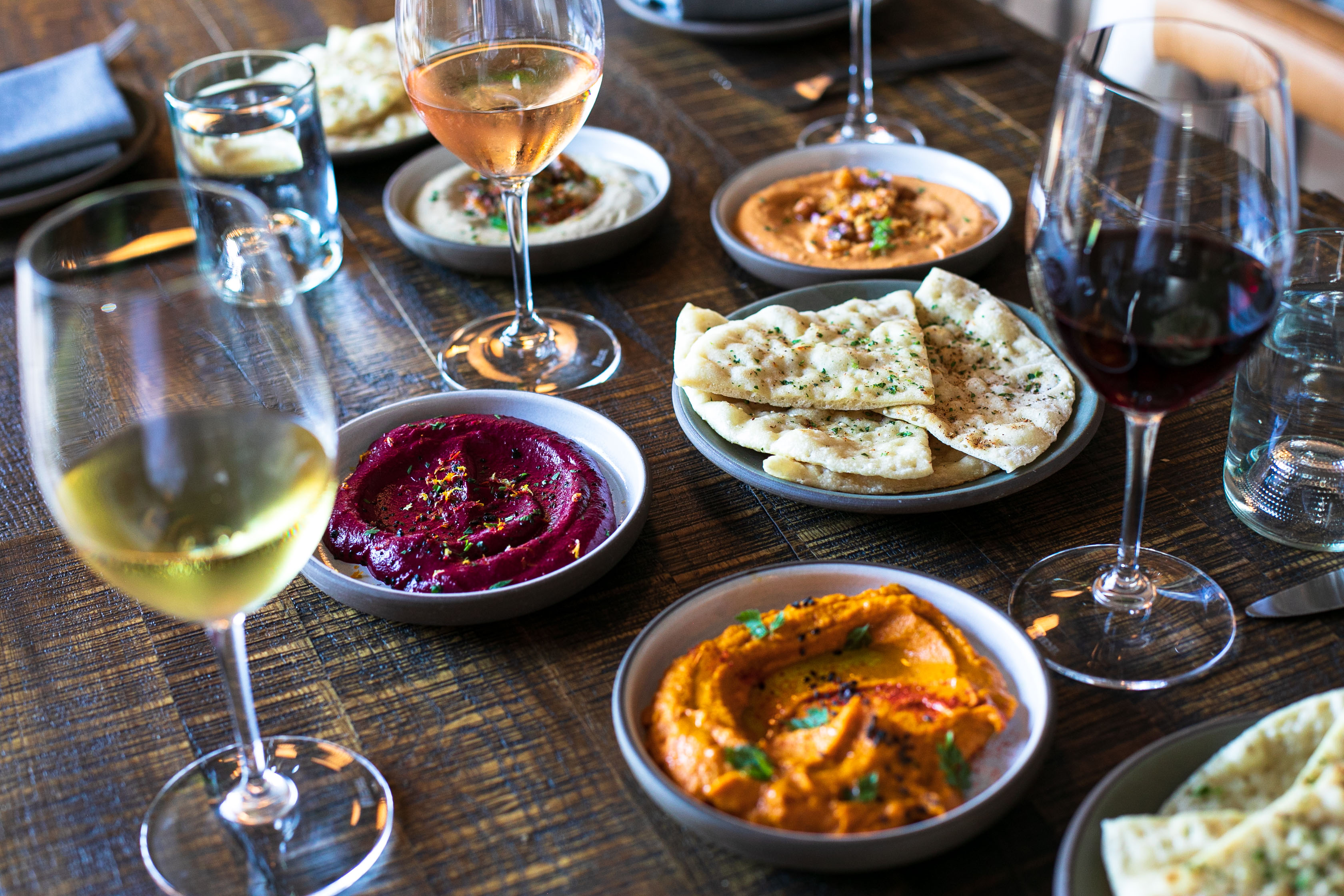 A dark wooden table set with a spread of various dips, pita, and wine