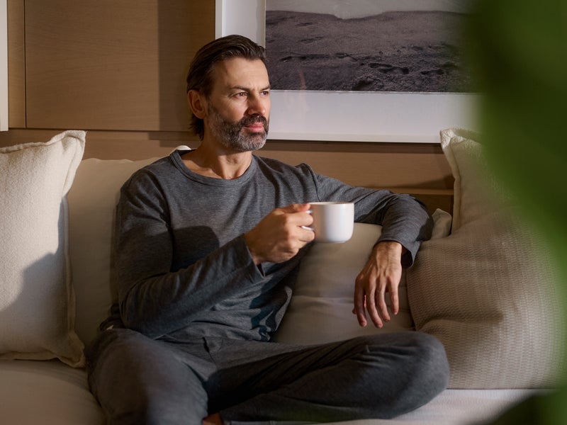 A man in loungewear sits on a plush couch drinking a coffee