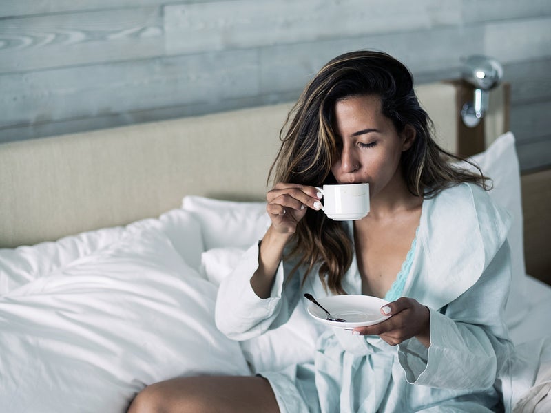 Person in a robe sitting on a bed sipping from a mug