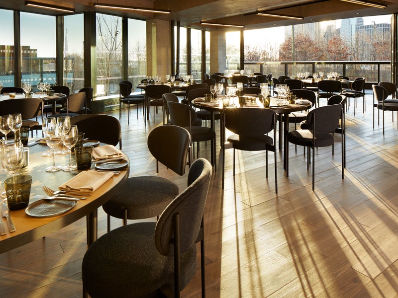 Aster Social Evening. A spacious room with large round wooden tables each set for eight individuals. Sunlight pours in through the windows.