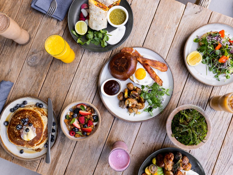 A full table spread of various breakfast dishes, including from left to right, a stack of blueberry pancakes, a breakfast wrap, bacon and eggs, and a salad.