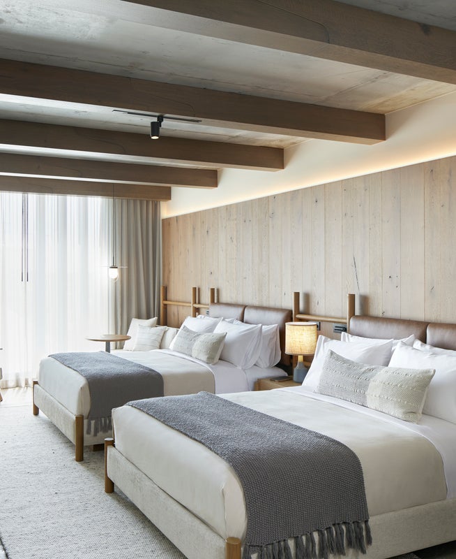 Two queen size beds with wooden headboards sit side by side basking in the sun shining through the nearby window