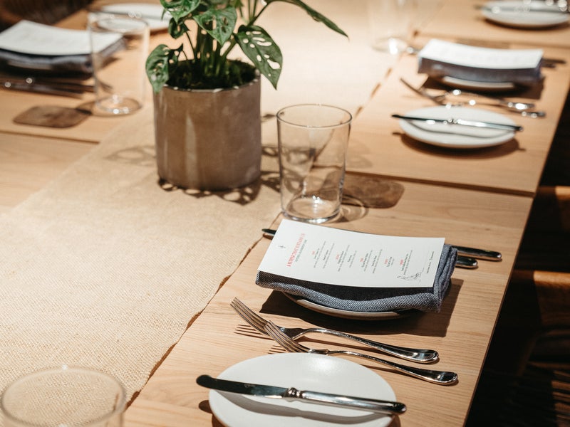 A long wooden banquet table with elegant seating placements.  A beige runner is placed lengthwise down the middle, accented with plants.