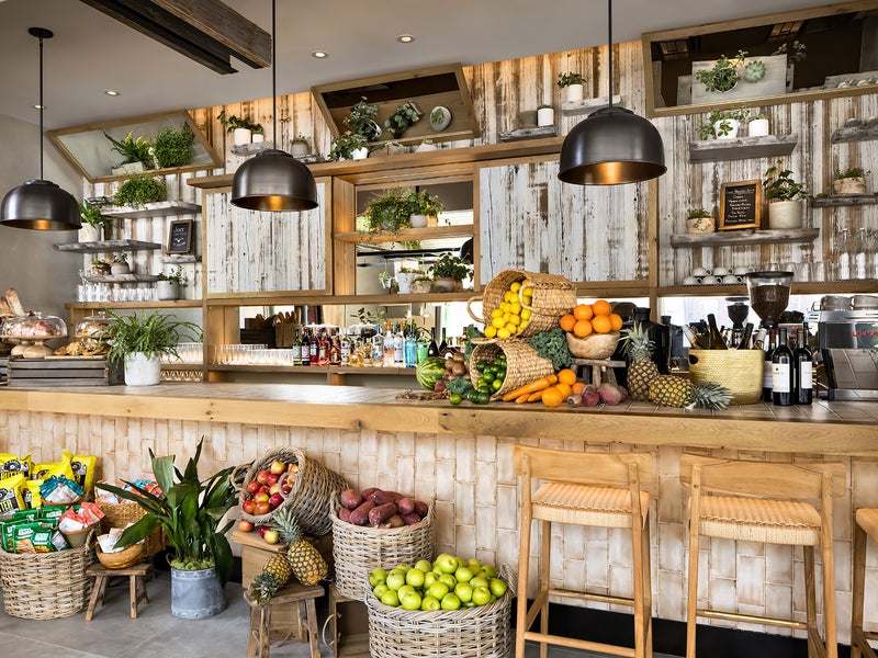 Baskets of fruit decorate a lengthwise wooden bar with stool seating.  Displays of drinks, coffee machines, dishes, and food can be seen lining the wall behind the bar.
