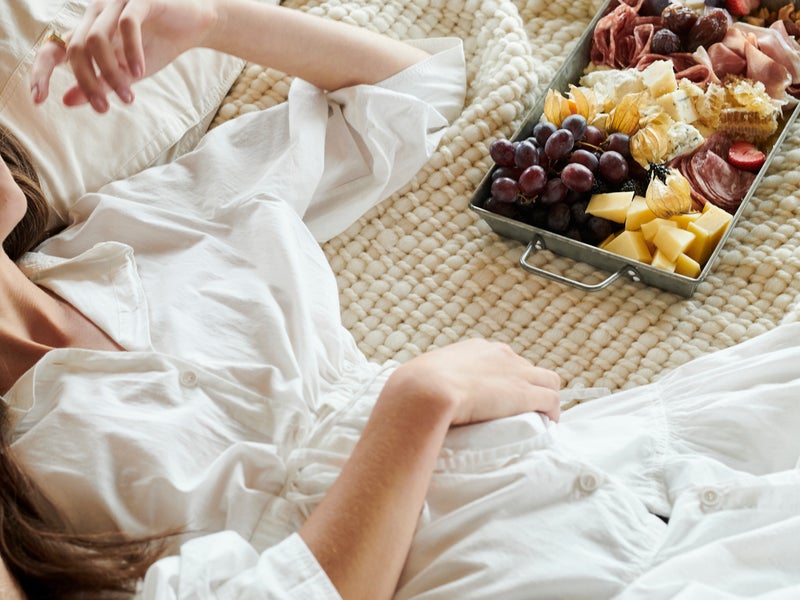 A woman in a white robe lounges on her bed next to a charcuterie plate