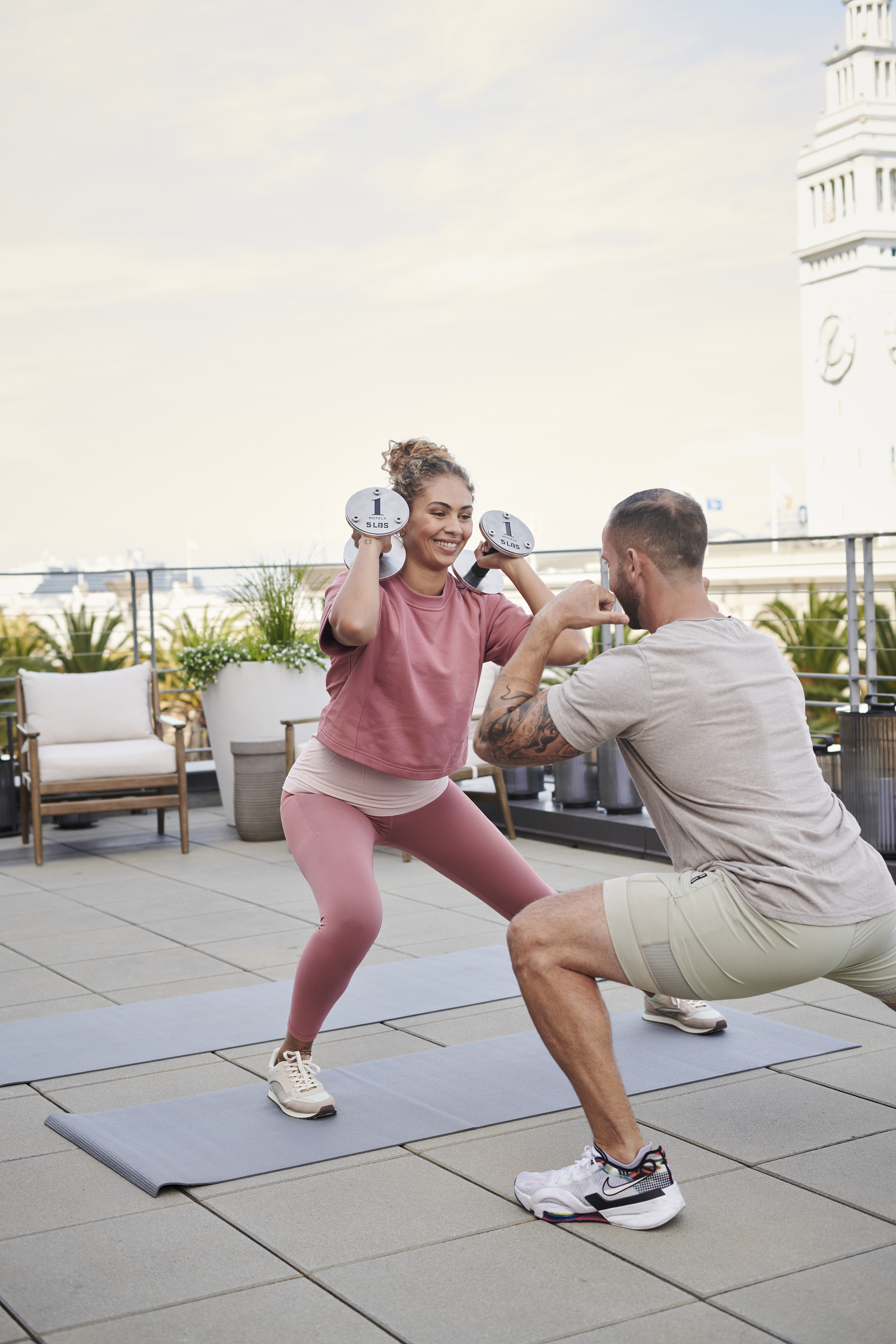 Un istruttore privato di sesso maschile istruisce una cliente di sesso femminile che, tenendo in mano dei pesi liberi, esegue uno squat laterale.