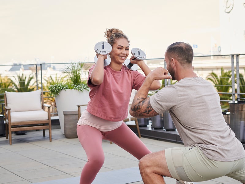 A male private trainer instructs a female client holding free weights, performing a side squat