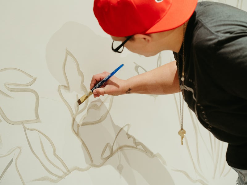 An artist with a backwards orange ball cap holds their brush steady as they paint an outline of flowers on a white wall