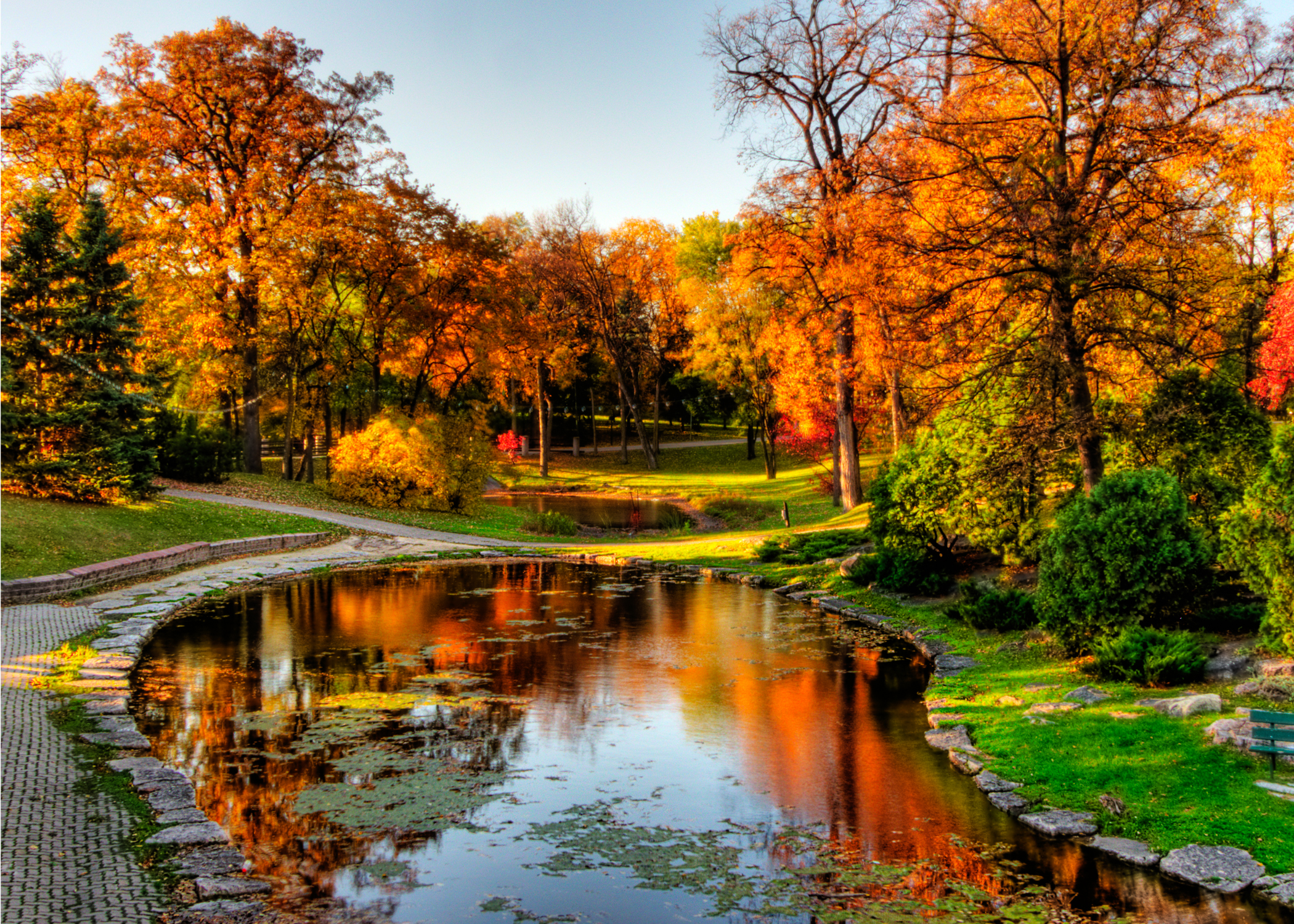 Sun shining over a small lake, walking trail and fall colours in Kildonan Park