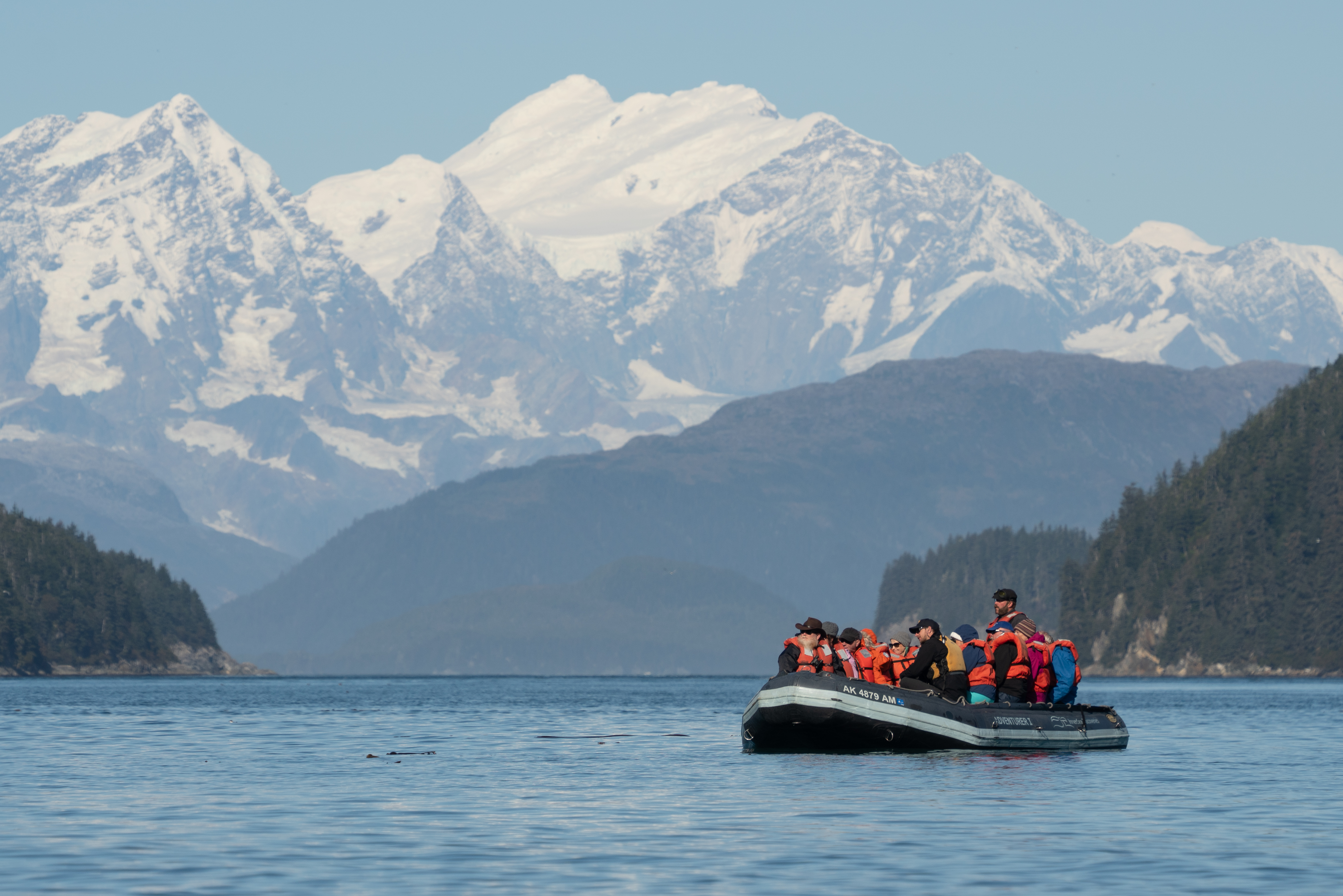 Group of guests ride in a zodiac boat through an inlet in Alaska with mountains and forest in the background.