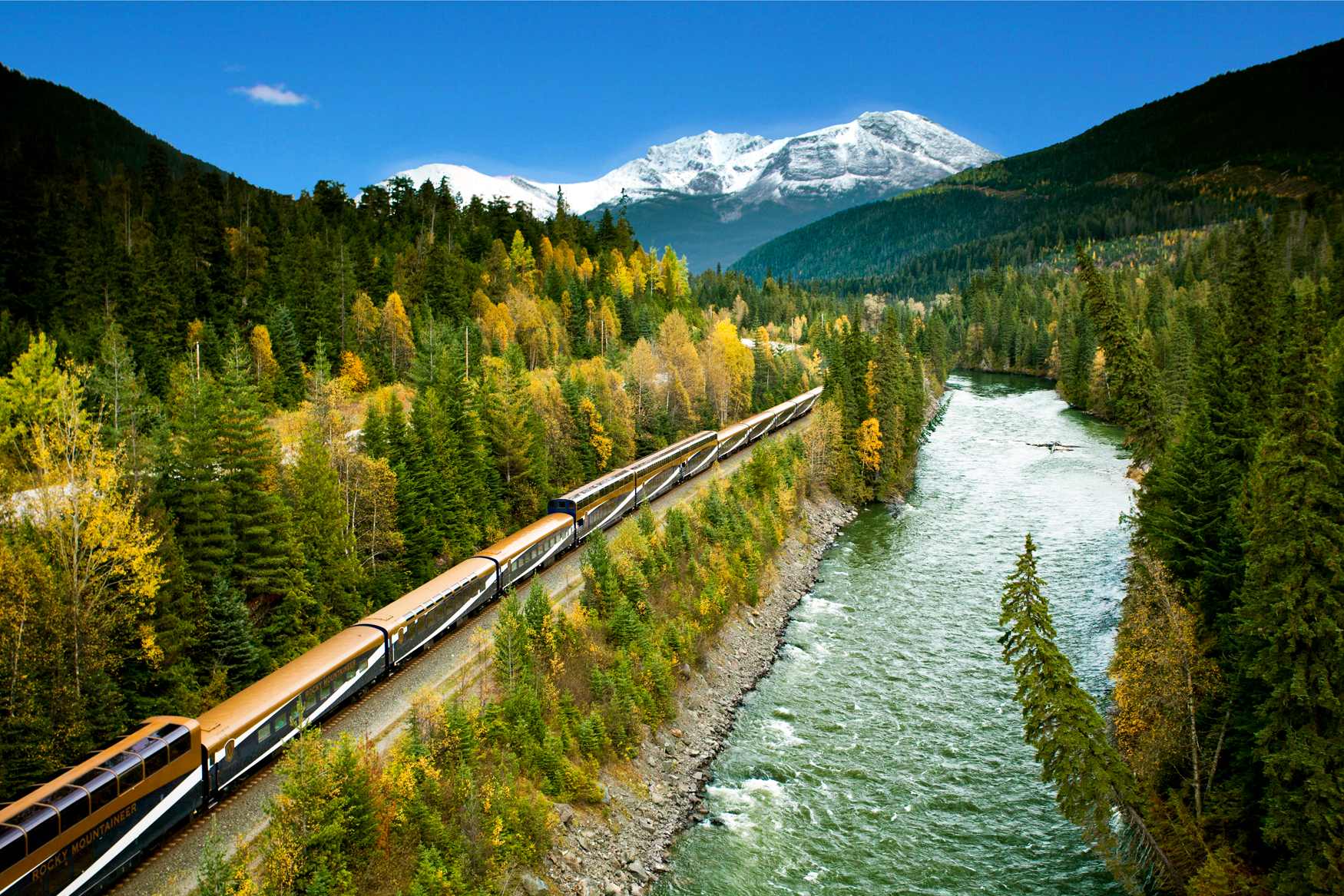 Rocky Mountaineer rides along river and forestry with Canadian Rockies in the background during fall