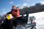 Senior woman wearing puffer jacket smiles while sitting on snowmobile on fresh snow with mountains behind her