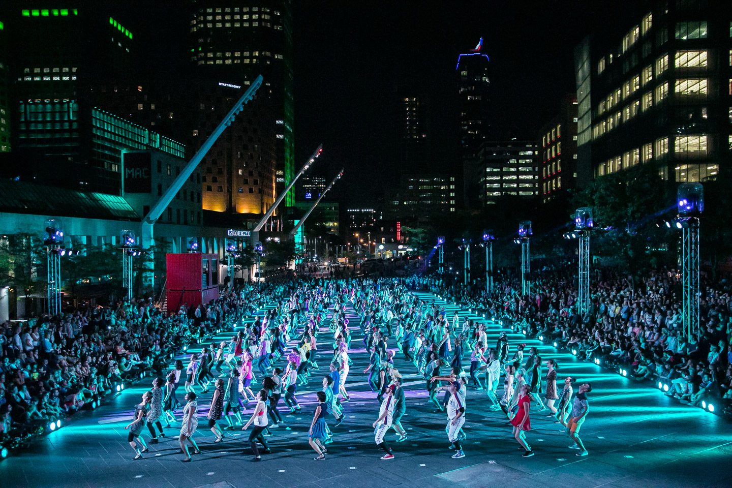 Large group of dances performing in a plaza at night as a crowd watches