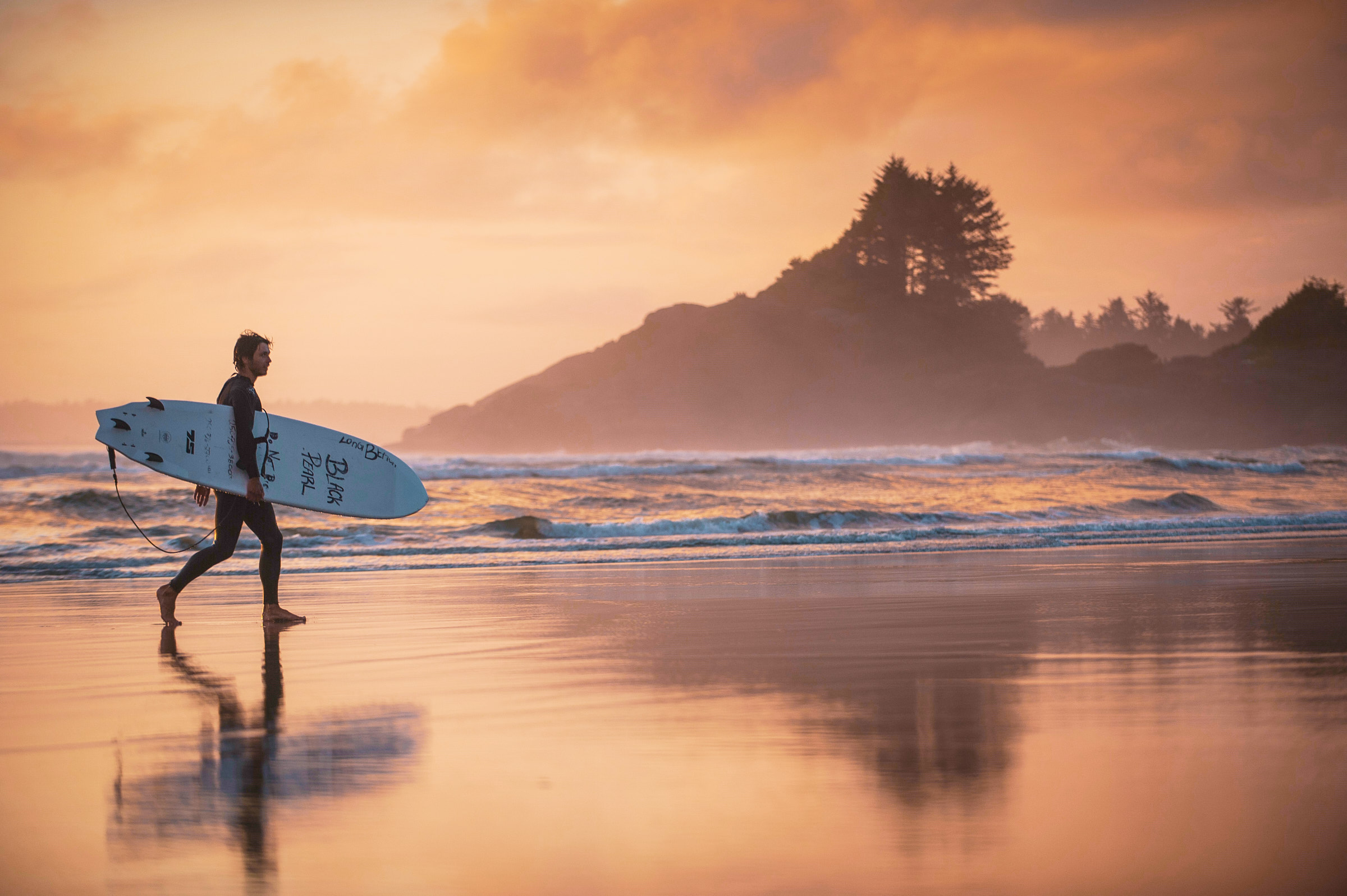 A surfer is walks along the beach at sunset in Tofino with surfboard under their arm