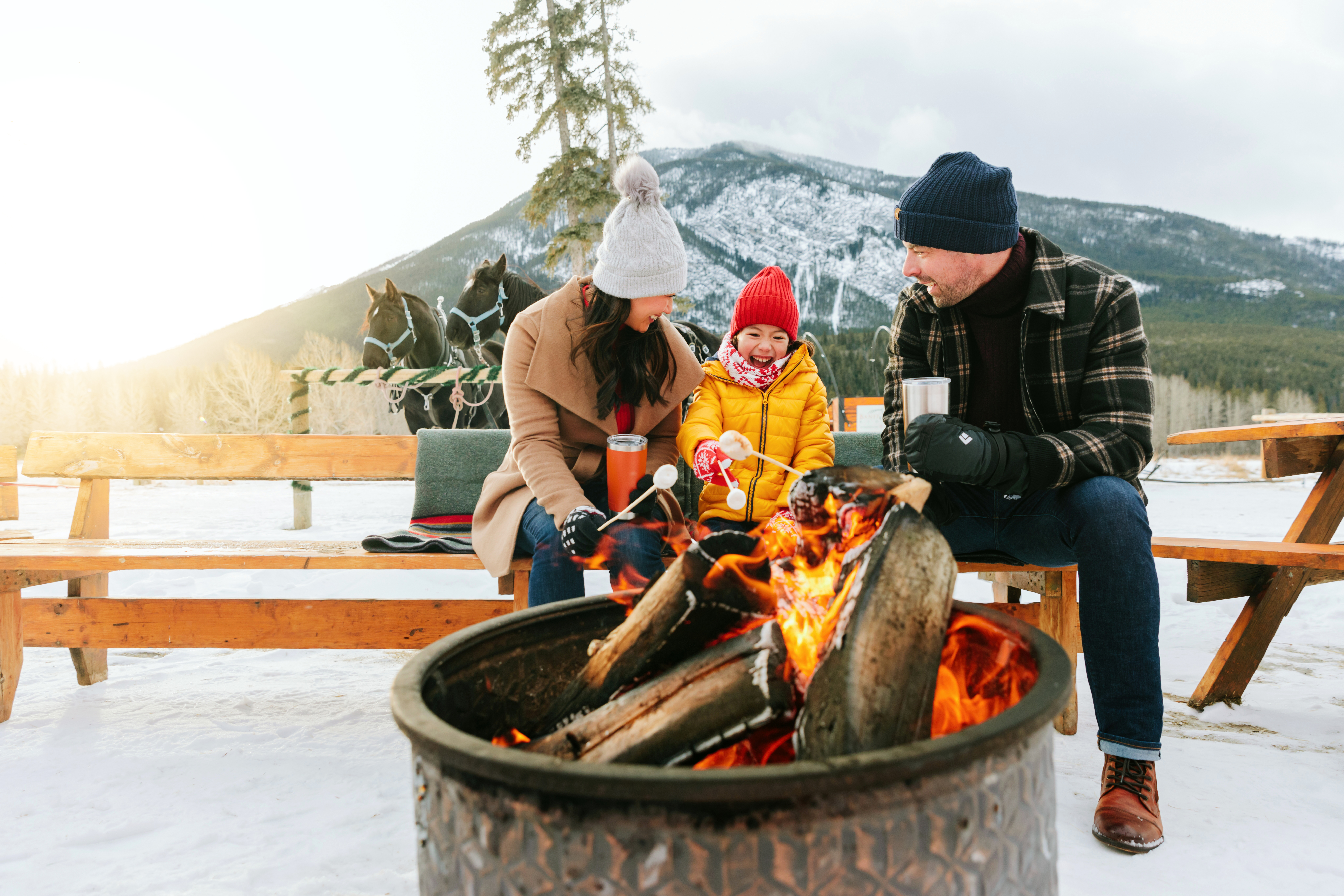 A family roasts marshmallows over fire while sitting on outdoor bench with a mountain in the background