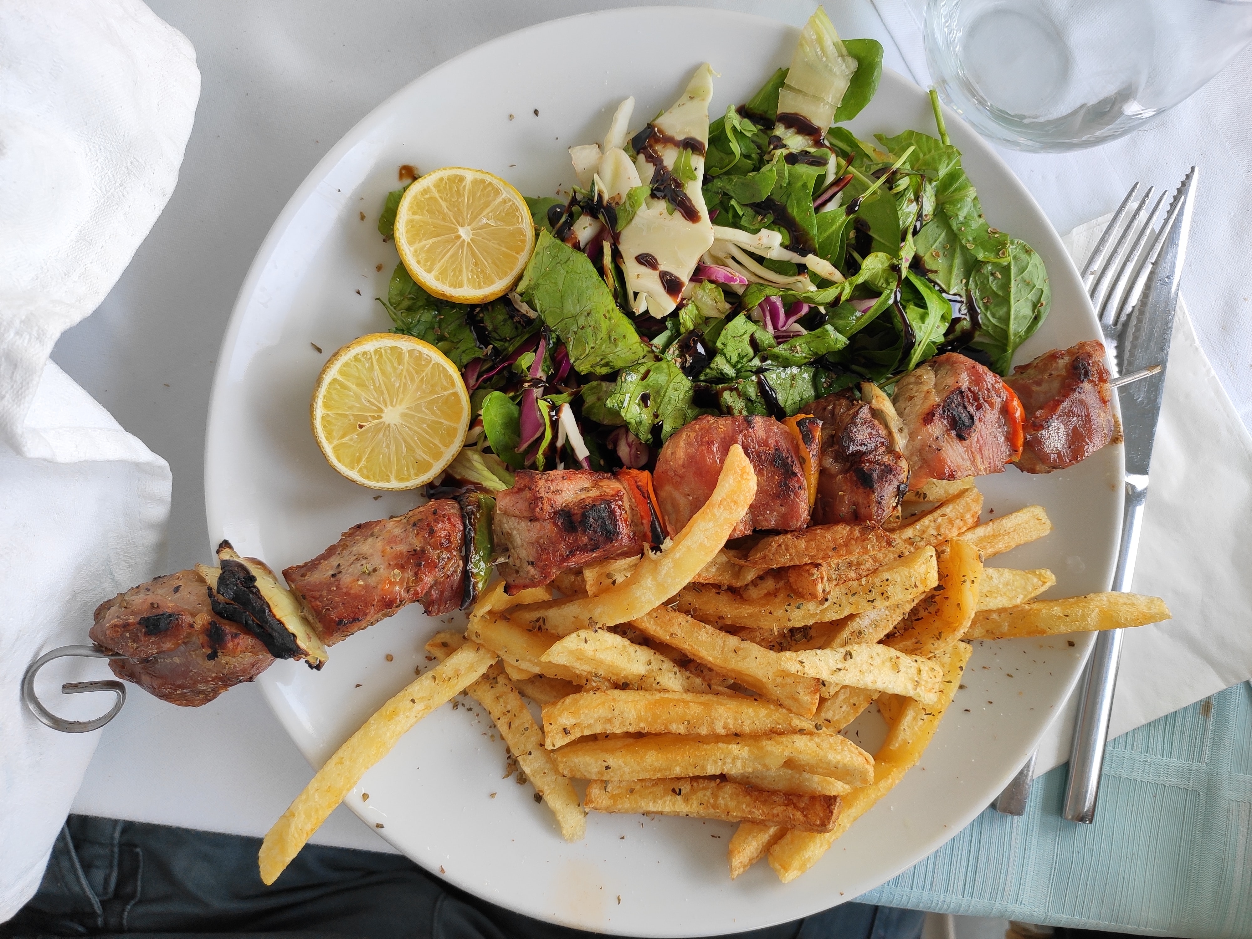 Serving of Souvlaki with meat skewer, French fries, lemon and dressed salad on a plate