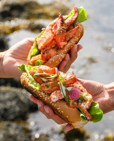 Hands holding two lobster roll sandwiches by the seaside