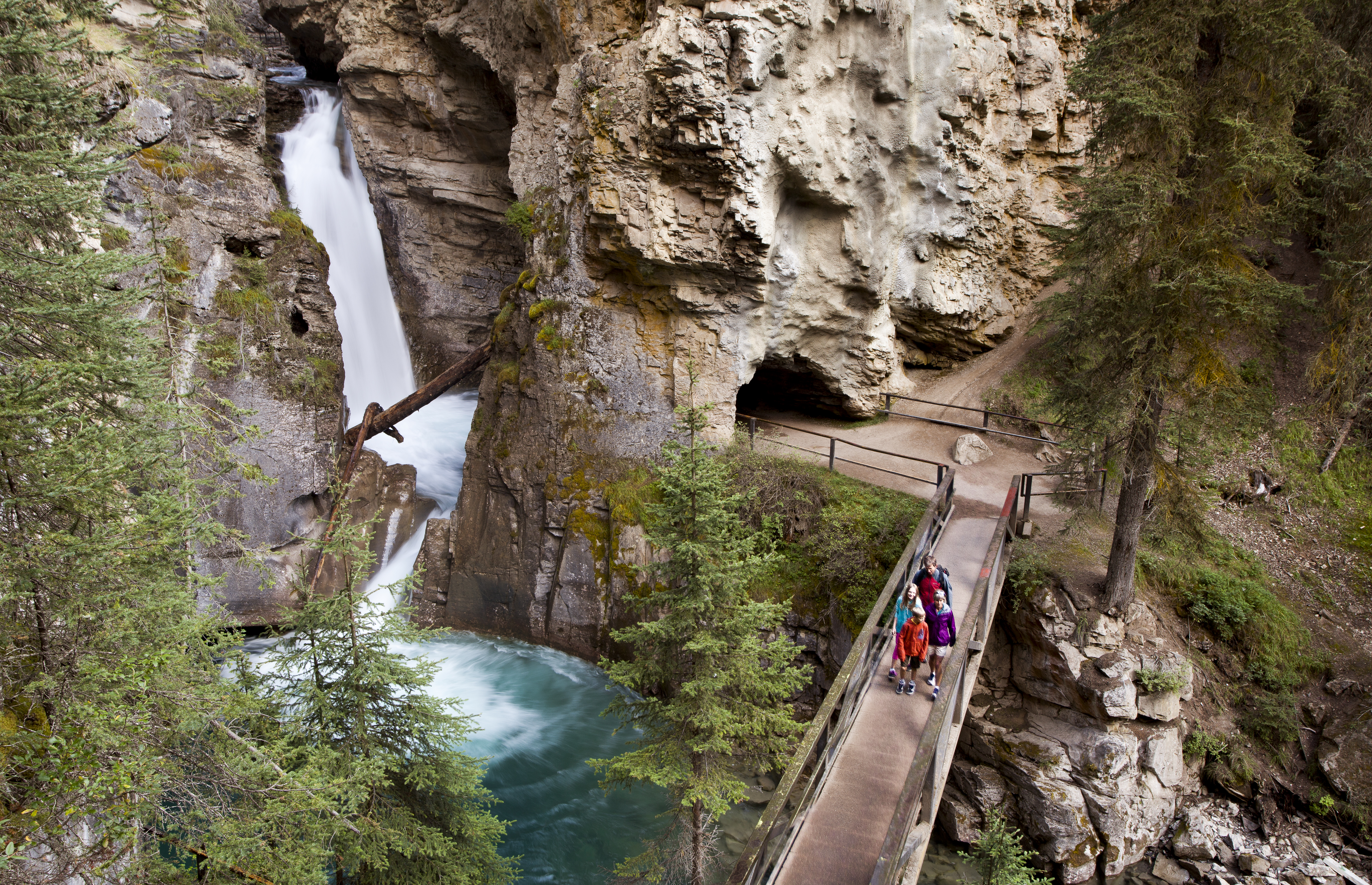 Family on catwalk cross waterfall rushing from cliff and into turquoise plunge pool in Johnston Canyon