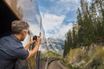 A man leans out of the Rocky Mountaineer train outdoor viewing area to take a photo of the mountains