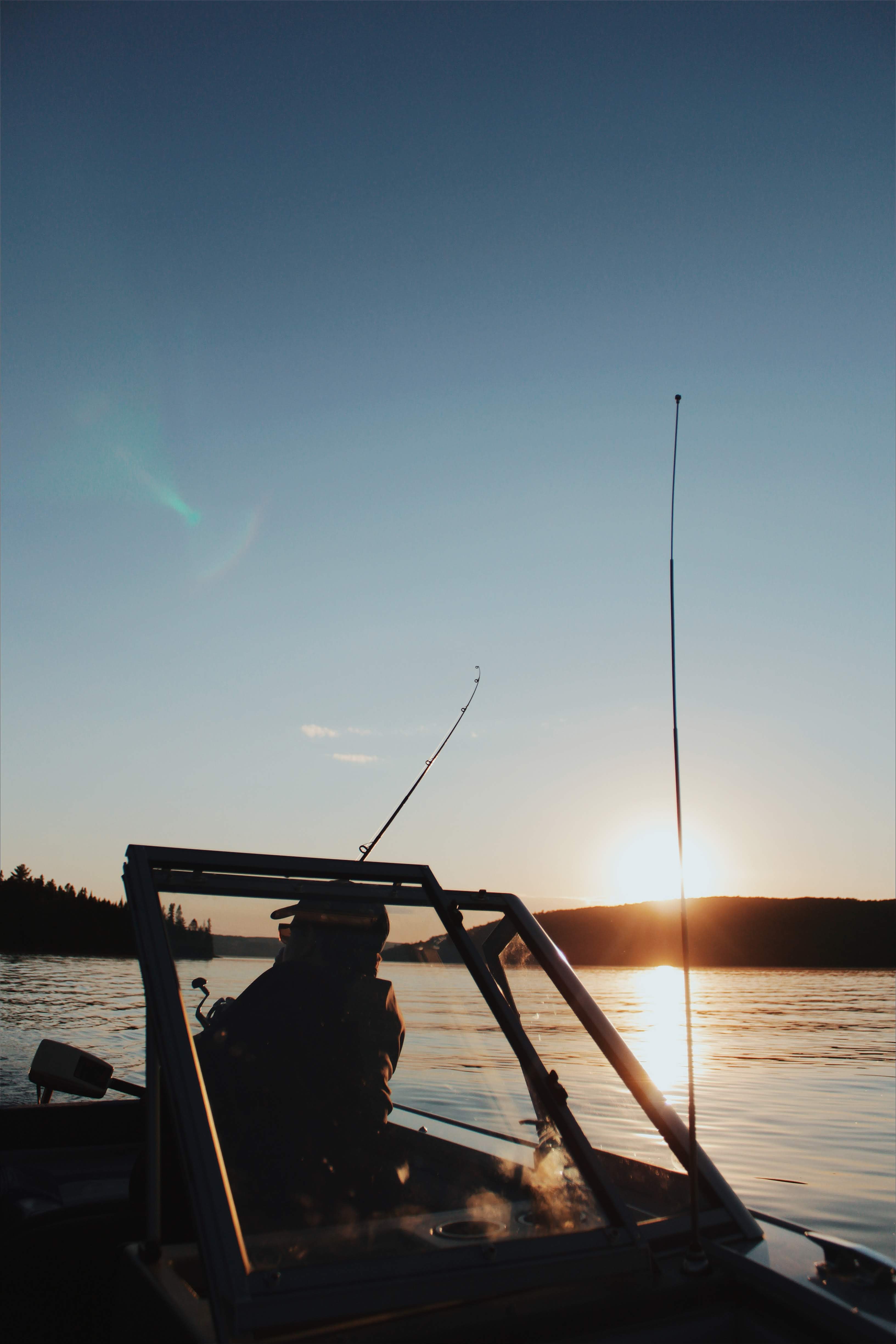 Person on boat with, fishing equipment, looks out towards shore landscape in the Bas-Saint-Laurent region while sun sets