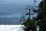 People sitting on a ski chair lift above the snow with Vancouver city and ocean in the distance