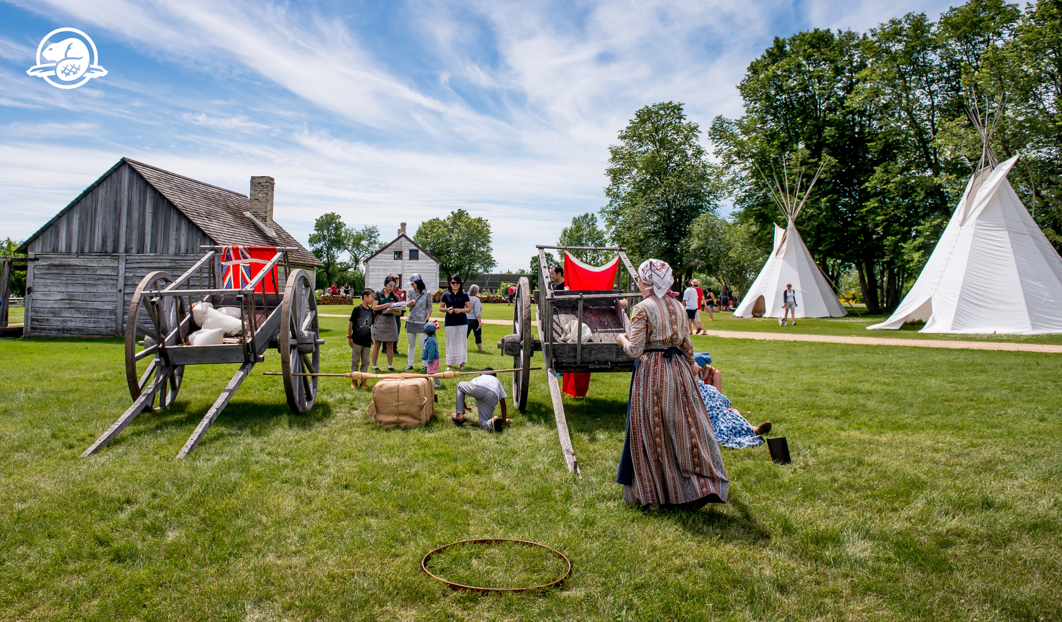 Person in costume standing in front of wooden wagons at a historical village 
