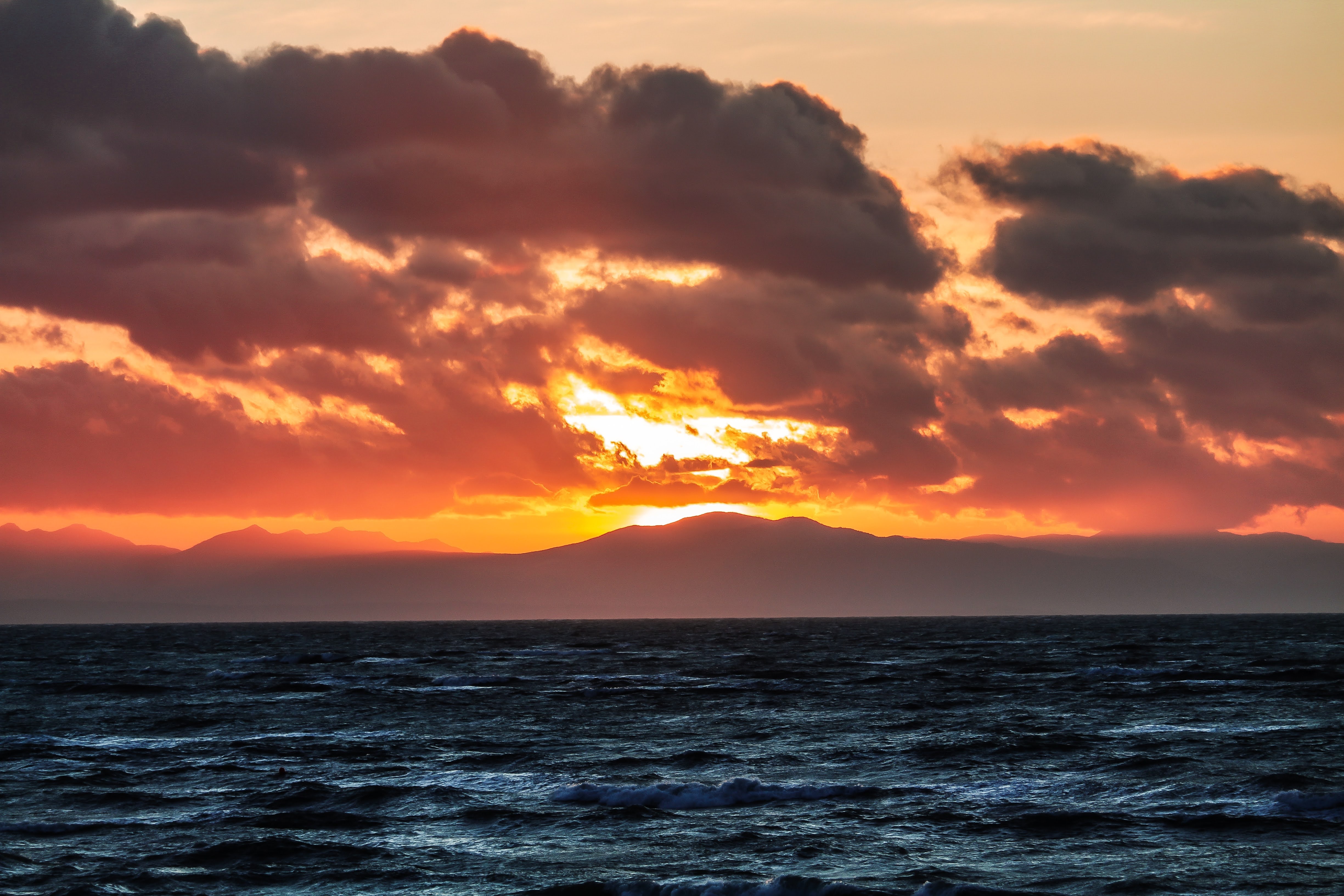 View of beach waves and horizon with sun setting behind mountains in the distance