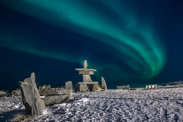 Inukshuk and visible aurora natural light display shimmers in the sky at night in Churchill, Manitoba
