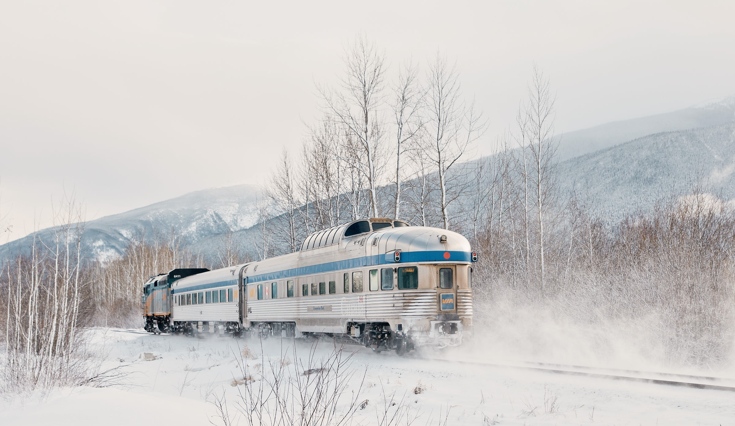 Silver train traveling through snowy landscape with snowy mountains in the background.