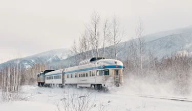 Silver train traveling through snowy landscape with snowy mountains in the background.